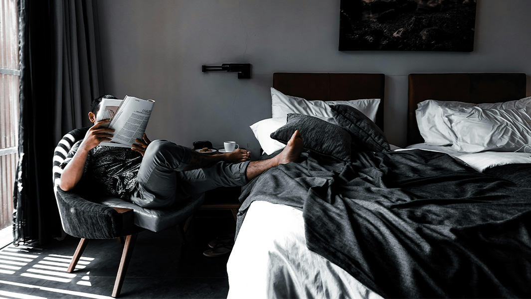 A black and white photo of a relaxed man reading on a chair with his feet on a bed