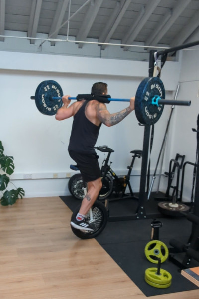 Jason facing away from the camera, on the unicycle, holding the barbell