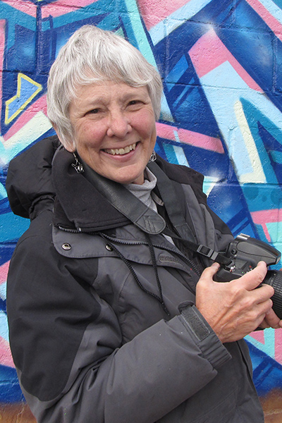 martha cooper holding a camera in front of a wall with graffiti