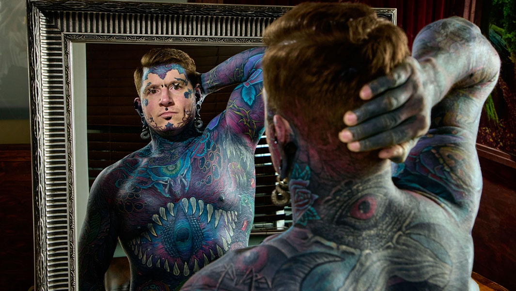 Tattooed man Remy holding one hand behind his head and looking in the mirror
