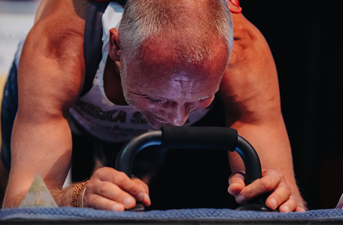 closeup of white haired man holding handlebars while performing an abdominal plank with a 40lb backpack