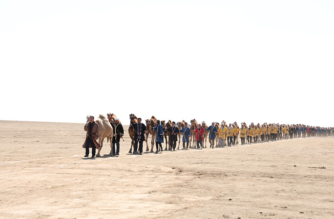 a row of camels are led by herdsman in a straight line parade