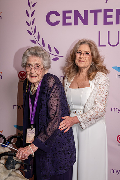 two elderly woman stand side by side in front of a purple backdrop