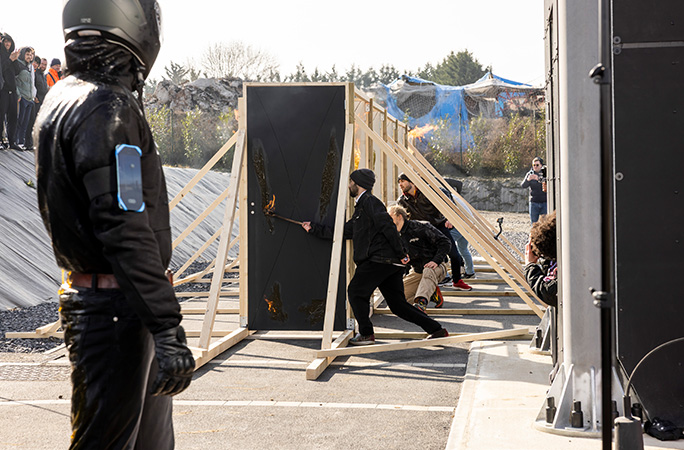 a man wearing a helmet stares at doors set in a row being set alight