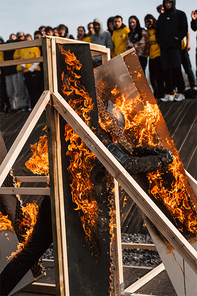 a man wearing a helmet and all black breaks through a door on fire while set on fire