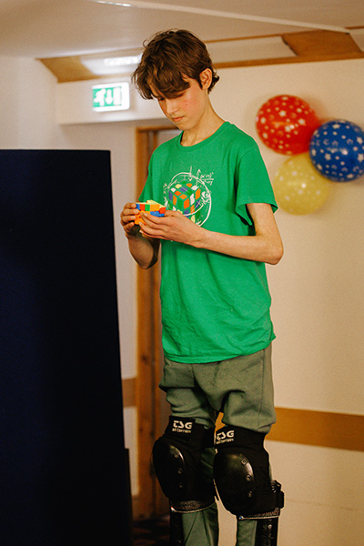 a boy wearing a green t shirt solves a puzzle cube while on stilts 