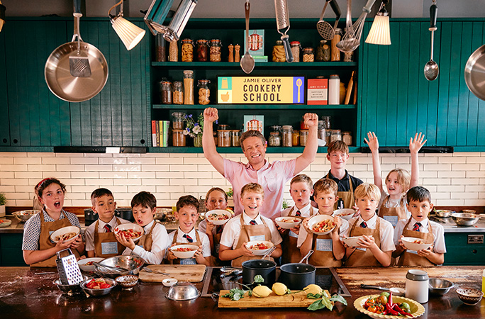 a man wearing a pink shirt cheers with children holding up bowls of pasta